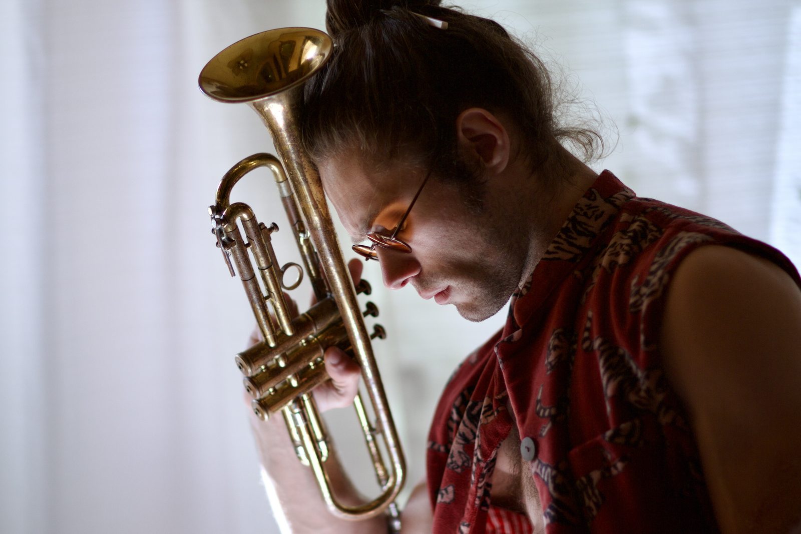 Jamen Krause holding a brass trumpet, head bowed, backlit against a bright window.